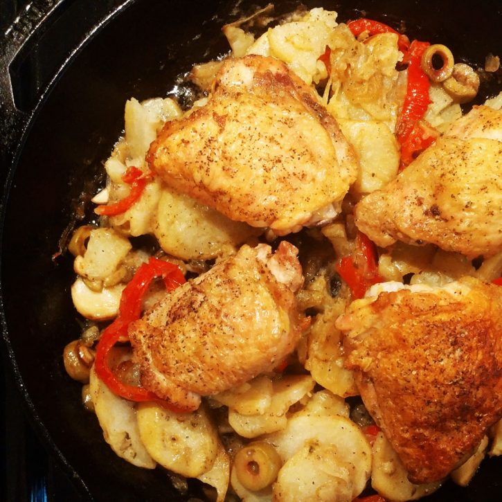 braised chicken in a cast iron pan with garlic, potatoes, olives